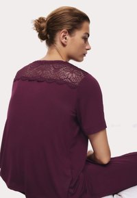 Top corto de manga corta morado con un panel de encaje en la parte posterior, que presenta un diseño de flores y una textura suave. Modelo mostrado de lado.