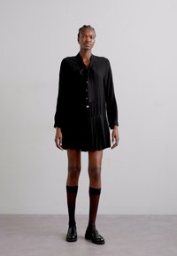 Robe noire à manches longues, patte de boutonnage à l'avant et nœud au niveau du cou ; détail de jupe plissée. Le mannequin porte des chaussettes au genou et des chaussures.