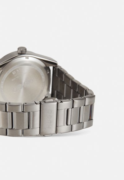 Montre en acier inoxydable avec un bracelet en métal poli. Présente un boîtier rond, un cadran argenté lisse et un fermoir gravé « HUGO ».