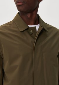 Olíva zöld vízálló dzseki, hegyes gallérral, gombos záródással és sima, matt textúrával. Látható varrások elöl.