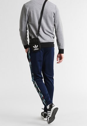 Sudadera gris con puños negros, pantalones de chándal azul marino con rayas multicolores a los lados, y bolso cruzado negro de Adidas.