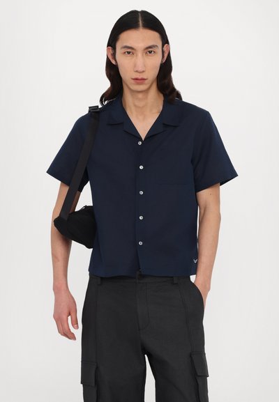 Marineblaues Kurzarmhemd mit Kragen, Knopfleiste und einer einzelnen Brusttasche; kombiniert mit schwarzen Cargo-Hosen und einer kleinen schwarzen Tasche.