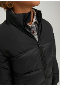 Osoba ubrana w czarną kurtkę puchową z zapięciem na suwak, noszącą pod spodem zapinaną na guziki koszulę w kratę, odsłaniającą część szyi i podbródka.