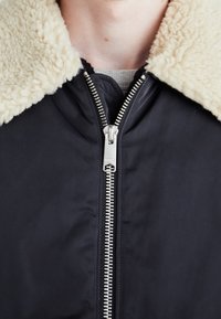 Chaqueta bomber negra con un cuello de piel sintética de color crema, cremallera frontal y tela texturizada. Diseño simple y funcional.