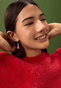 Guldtonede, træformede øreringe og en rød, fluffy sweater mod en grøn baggrund. øreringene har små dekorative detaljer.