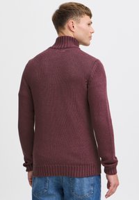 Maglione a maglia color bordeaux con colletto alto e polsini a coste, che mostra un motivo testurizzato. La vista posteriore rivela un orlo standard e una vestibilità rilassata.