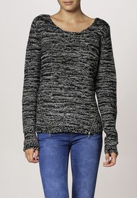 Schwarzer und weißer, strukturierter Strickpullover mit weitem Ausschnitt. Hat seitliche Reißverschlüsse und lange Ärmel. Wird über blauen Jeans getragen.