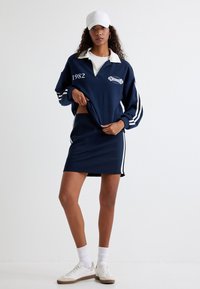 Felpa navy con colletto e strisce bianche, con "1982" e logo. Abbinata a una gonna navy con strisce laterali bianche e sneakers.