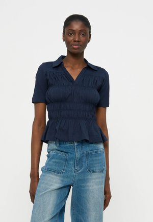 Moteris dėvi tamsiai mėlyną suraukytą trumpomis rankovėmis palaidinę su apykakle ir šviesiai mėlynas aukšto liemens džinsus su didelėmis priekinėmis kišenėmis.