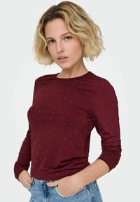 Blondynka z krótkimi, falowanymi włosami, ubrana w bordową bluzkę z długim rękawem i małymi białymi kropkami oraz jasnoniebieskie dżinsy, z rękami w kieszeniach.