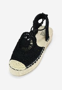 Sandalia de ganchillo negra con suela de yute trenzado. Presenta un diseño abierto con un acento decorativo redondo, correa ajustable y una puntera beige en contraste.