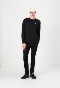Sudadera negra con puños acanalados, pequeño logo en el pecho, combinada con jeans ajustados negros y zapatillas negras. Una gorra neutra completa el conjunto.