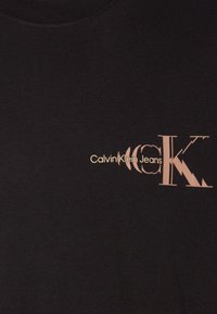 Černé bavlněné tričko s potiskem loga ve světle hnědé barvě. Design zahrnuje stylizovaný text s charakteristickým písmem a akcenty.