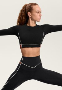 Fekete sportos crop top fehér és barna díszítésekkel, hosszú ujjakkal és testhez simuló kialakítással, magas derekú leggingsszel párosítva.