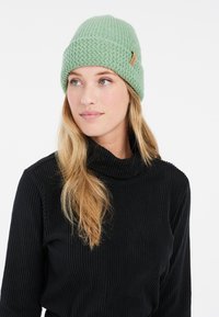 Hellgrüne gestrickte Mütze mit gerippter Textur und einem braunen Lederetikett. Getragen mit einem schwarzen, gerippten Rollkragenpullover, bei dem lange Haare sichtbar sind.