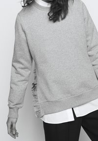 Hellgrauer Sweatshirt mit gerippten Bündchen und Saum, versehen mit strukturierten Rüschen an den Seiten. Wird über einem weißen Hemd mit Kragen getragen.
