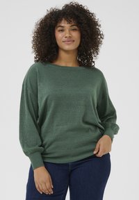 Grön stickad pullover med vid halsringning och långa, lösa ärmar. Tyget har en lätt lyster, kompletterad med tighta manschetter och nederkant.