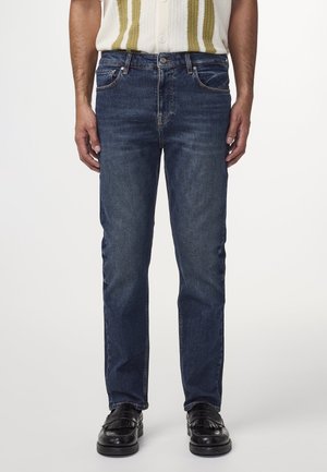 Mand iført mørkeblå straight-fit jeans, sorte læder loafers og en hvid strikkevest med grønne lodrette striber.