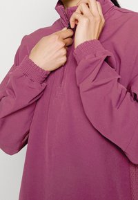 Jersey morado con cuello de media cremallera, puños elásticos y tejido texturizado, con un corte holgado y detalles de costura mínimos.