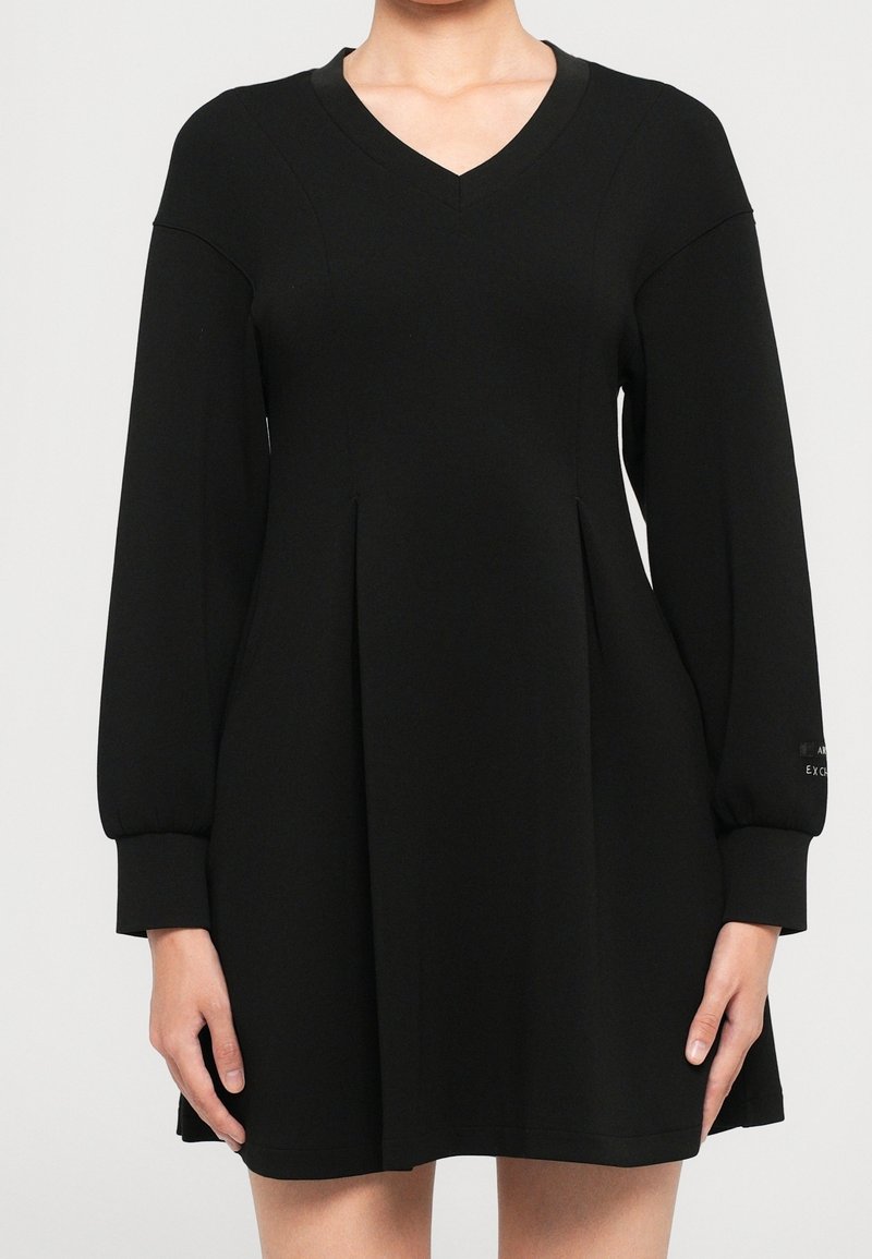 Robe noire à manches longues avec décolleté en V et plis, portée par une personne visible de la poitrine jusqu'à mi-cuisse, les mains détendues sur les côtés.