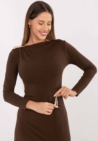 Brązowa sukienka z długim rękawem wykonana z rozciągliwego materiału, z drapowanym dekoltem i złotym akcentem na ramieniu, z marszczeniem po bokach.