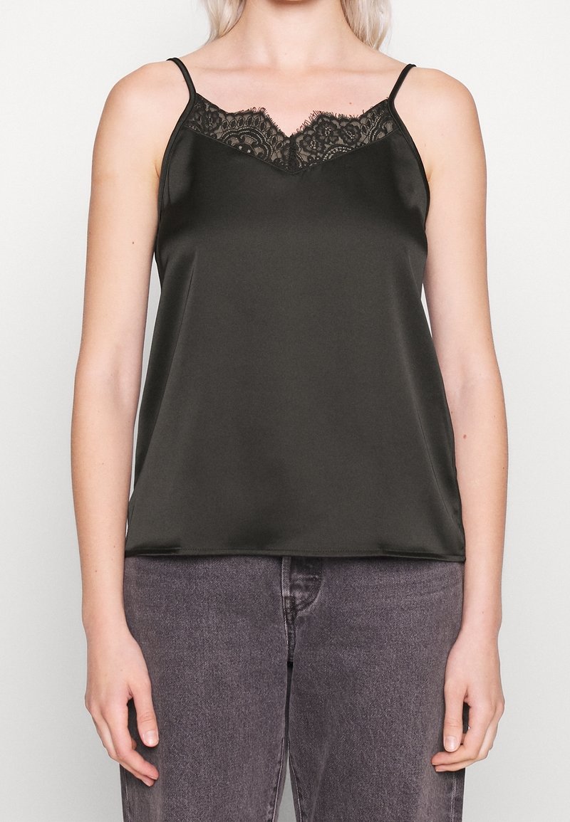 Camisole en satin noir avec bordure en dentelle au niveau de l'encolure, portée avec un jean en denim noir délavé sur une personne à la peau claire et aux cheveux blonds.
