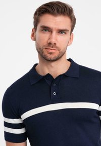 Mörkblå polo skjorta i stickad tyg med ett vitt horisontellt rand och två vita detaljer på ärmarna, avslutad med en tre-knapps ståndkrage.