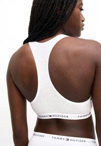 Valge spordirinnahoidja racerback-disainiga, pehme kangas ja bränditud elastne rihm. Seljal on suur lõhik paremaks õhutusvõimeks.