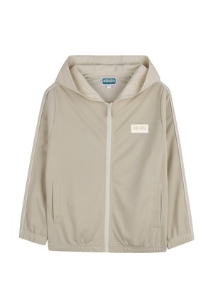 Beige jas met rits en capuchon, gemaakt van gladde stof. Voorzien van witte strepen aan de zijkant en een logopatch op de linkerborst. Twee voorkanten zakken.