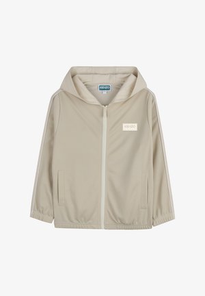 Veste beige zippée avec capuche, en tissu lisse. Présente des bandes blanches sur les côtés et un écusson logo sur la poitrine gauche. Deux poches avant.