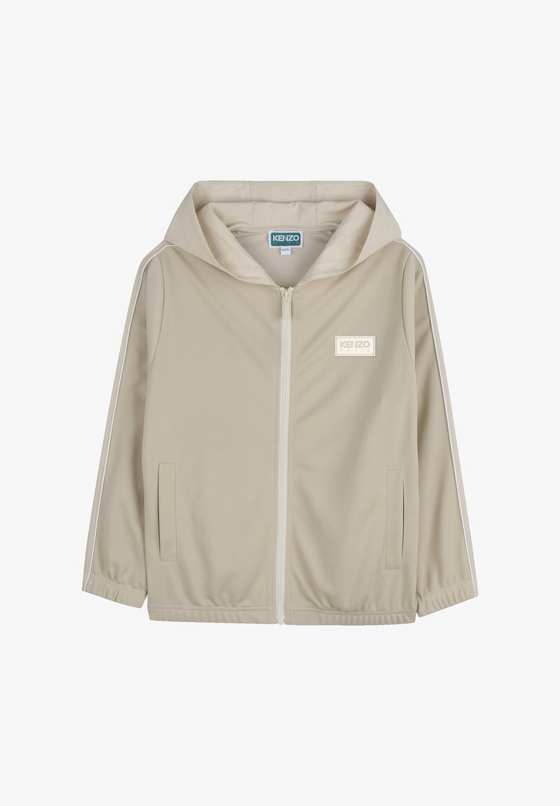 Beige jas met rits en capuchon, gemaakt van gladde stof. Voorzien van witte strepen aan de zijkant en een logopatch op de linkerborst. Twee voorkanten zakken.