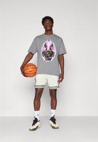 Grå grafisk t-shirt med et farverigt ansigtsdesign, hvide mesh-shorts med sorte striber, og sneakers med sorte og gyldne accenter.