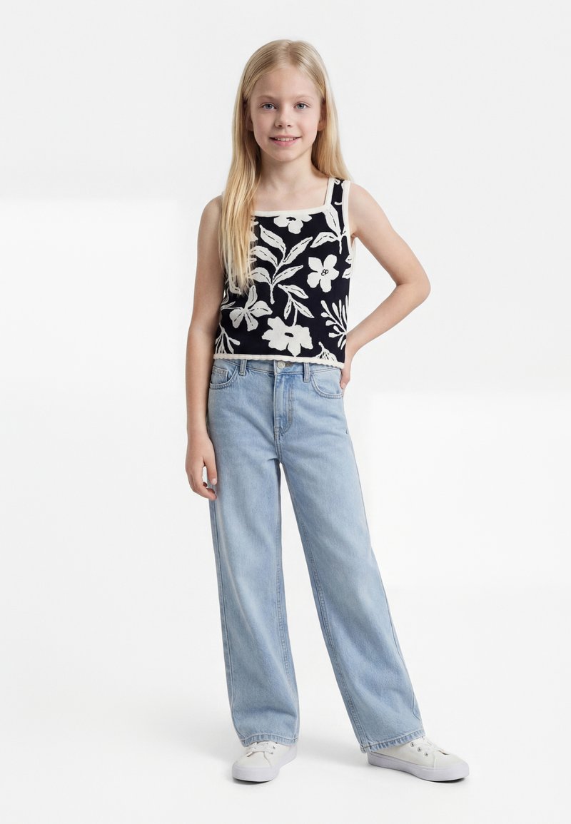 Nuori tyttö, jolla on pitkät vaaleat hiukset ja joka käyttää mustavalkoista kukallista toppia, vaaleansinisiä leveälahkeisia farkkuja ja valkoisia tennareita, seisoo käsivarret lanteilla.