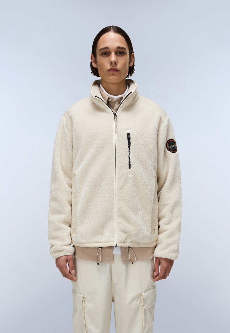 Jachetă din polar de culoare bej deschis, cu fermoar în față, guler înalt și un buzunar pe pieptul stâng. Are un patch rotund cu logo pe mâneca dreaptă.
