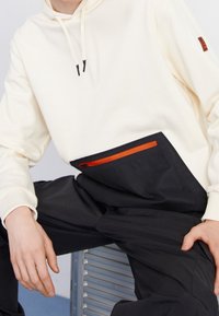 Sudadera crema con acentos negros y un bolsillo con cremallera naranja en contraste; combinada con pantalones negros, creando un look en capas con tejido texturizado.