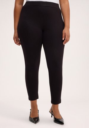 Kvinde iført sorte, tætsiddende ankellange bukser og sorte stiletter med spids tå og slingback, stående mod en ensfarvet baggrund.