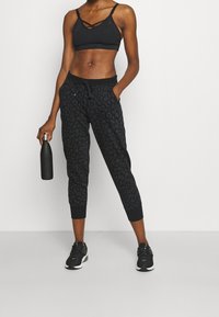 Pantalones deportivos con estampado de leopardo negro, puños elásticos y cintura con cordón, combinados con un sujetador deportivo negro. La modelo sostiene una botella de agua negra.