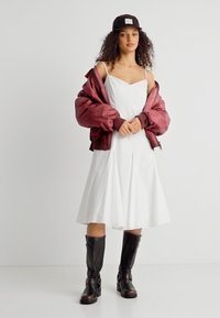 Γυναίκα που φοράει λευκό φόρεμα χωρίς μανίκια, μαύρο μπουφάν μπορντό ριγμένο πάνω στους ώμους, μαύρο καπέλο και μαύρες δερμάτινες μπότες μέχρι το γόνατο, στέκεται μπροστά σε λευκό τοίχο.