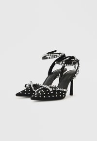 Melni augstpapēžu kurpes ar smailiem purngaliem, rotātas ar sudraba kniedēm un loku priekšpusē, ar potīšu siksniņām un spīdīgu apdari.