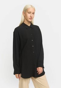 Musthave musta pikkade varrukatega nööbitav särk kerge kangaga, millel on krae, lahti lõige ja nähtavad nööbid eest.