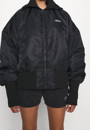 Osoba nosząca czarną kurtkę bomber Adidas na zamek z prążkowanymi mankietami i dołem, w połączeniu z czarnymi teksturowanymi szortami.
