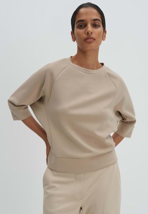 Frau steht mit Händen in der Hüfte, trägt ein beiges Dreiviertelarm-Oberteil und passende Hose vor einfachem hellem Hintergrund.