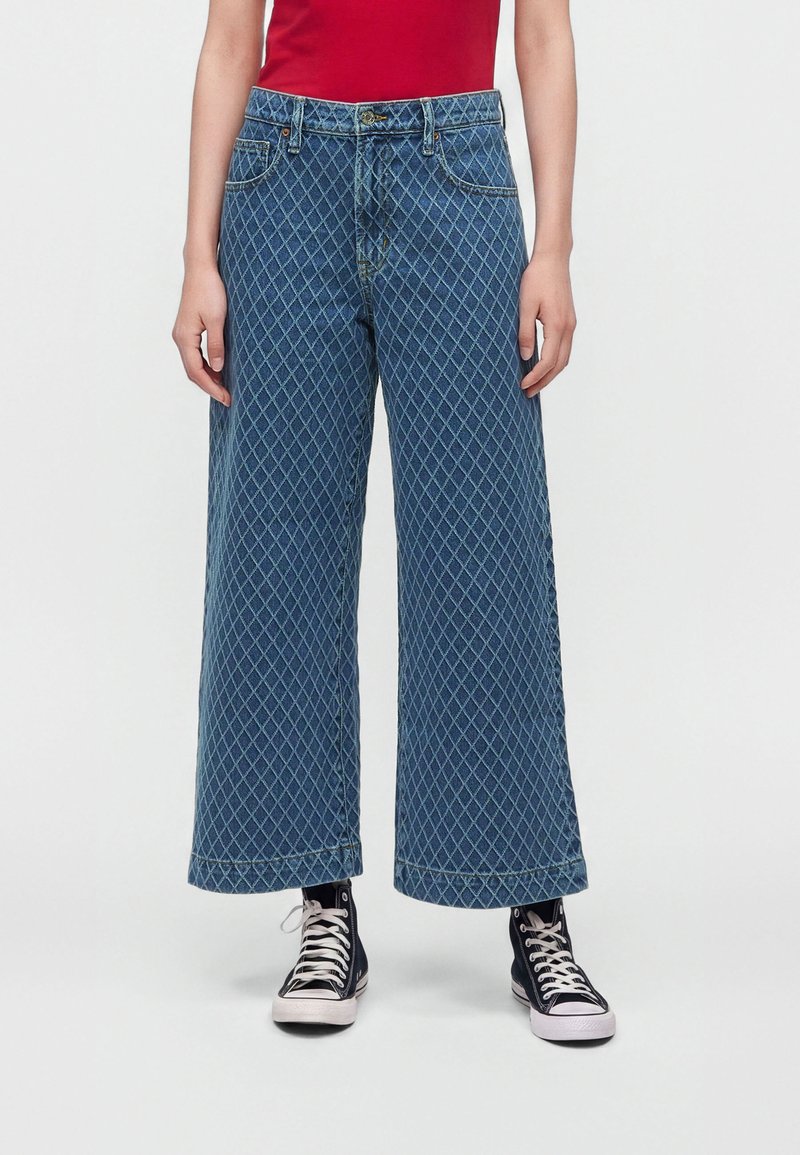 Personne portant un jean bleu large à motif losange, un haut rouge et des baskets montantes noires, debout devant un fond uni.