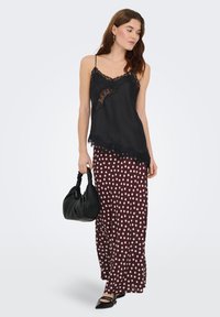 Camisole en satin noir avec bordure en dentelle, associée à une jupe longue bordeaux à pois. Le mannequin tient un sac à main noir texturé.