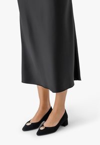 Žena v černé sukně do poloviny lýtek a černých botách na blokovém podpatku s velkými kulatými kovovými ozdobami nahoře.