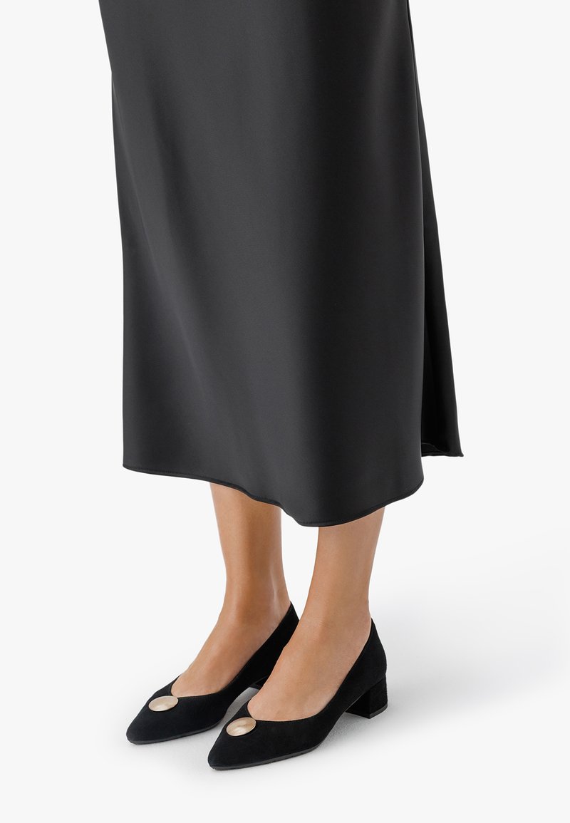 Žena v černé sukně do poloviny lýtek a černých botách na blokovém podpatku s velkými kulatými kovovými ozdobami nahoře.