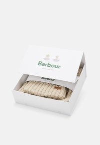 Béžová pletená čepice uvnitř bílé krabičky se zeleným písmem. Krabička má logo a název značky "Barbour." Čepice má texturovaný vzor a štítek.