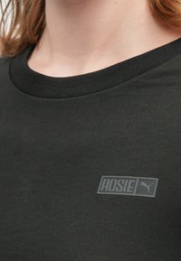 Czarna bawełniana koszulka z okrągłym dekoltem z teksturowaną naszywką z logo "ROSIE" i ikoną pumy po lewej stronie. Gładka struktura materiału.