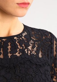 Zbliżenie na osobę w czarnej koronkowej bluzce z kwiecistym wzorem i okrągłym dekoltem, pokazującą część szyi i ust.