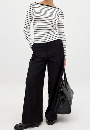 Woman wearing a white long-sleeve shirt with black stripes, black wide-leg pants, black loafers, and holding a large black handbag.